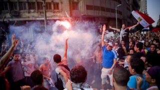 A crowd gathers in Tahrir Square in Cairo during the revolution in 2011; people raise hands and light flares.