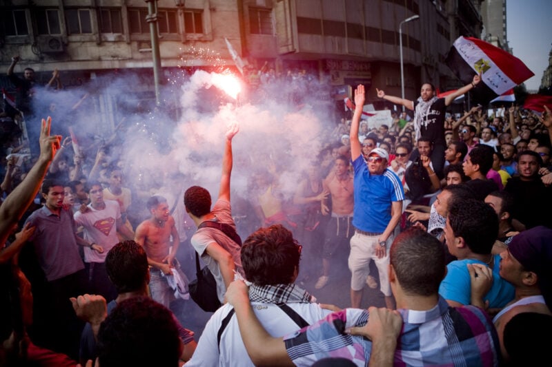 A crowd gathers in Tahrir Square in Cairo during the revolution in 2011; people raise hands and light flares.